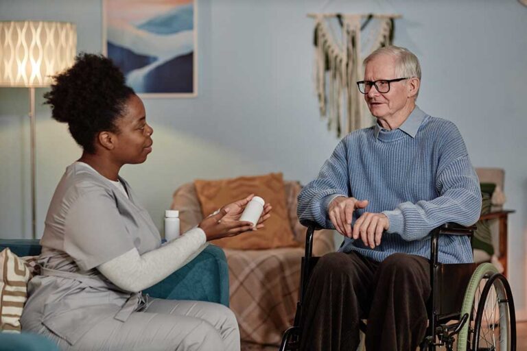 An older adult and a caregiver sharing a conversation in a sunlit assisted living common area.